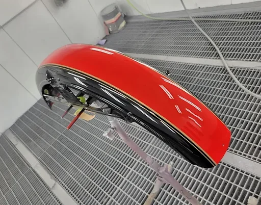 A newly painted red motorcycle fender with a black bottom and two thin gold pinstripes is in a paint booth.