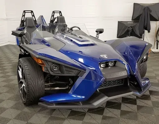 A blue Polaris Slingshot, a three-wheeled roadster, parked indoors on a black and grey tiled floor.