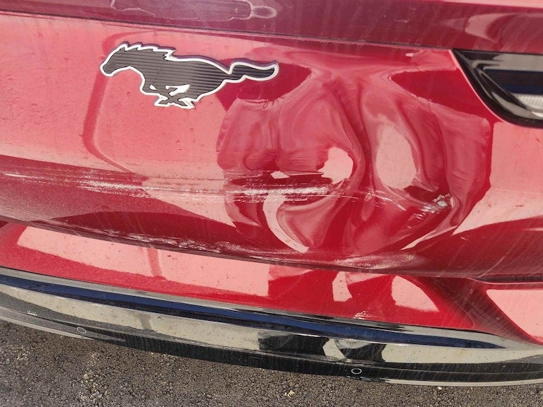 Rear view of a red Ford Mustang with a damaged and scratched bumper, visible denting, and silver scuff marks.