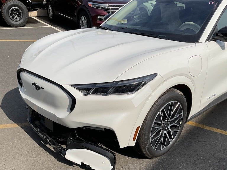 Front view of a white Ford Mustang Mach-E with its front bumper partially detached and hanging.