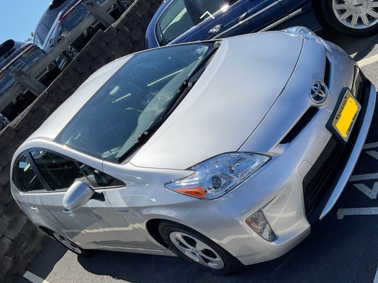 A silver Toyota Prius parked at an angle with a blue sedan and parked cars on a wooden structure in the background.