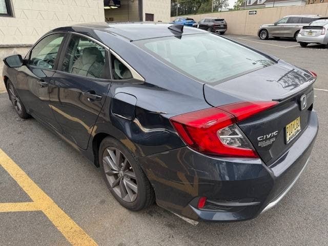 Rear 3/4 shot of a dark gray Honda Civic parked in a lot, with other cars and a building in the background.