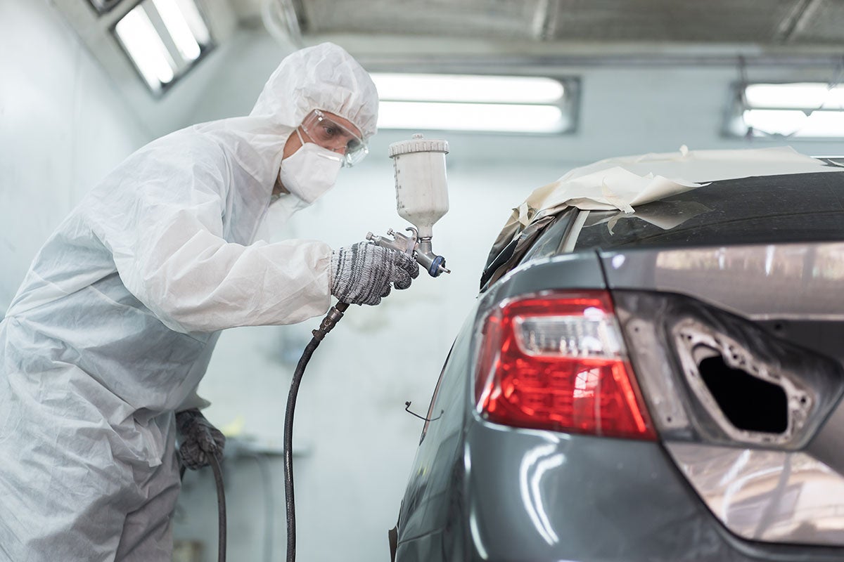 A person in a white hazmat suit, mask, and goggles spray painting the back of a grey car.